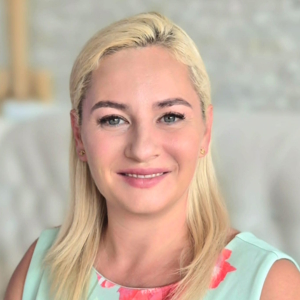 Uśmiechnięta kobieta o blond włosach w jasnej sukience z kwiatowym wzorem patrzy w obiektyw.