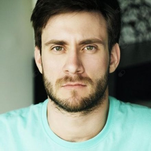 Mężczyzna o ciemnych włosach i brodzie w turkusowej koszulce patrzy w obiektyw.