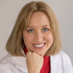 Uśmiechnięta kobieta o blond włosach w białym fartuchu medycznym opiera brodę na dłoni.