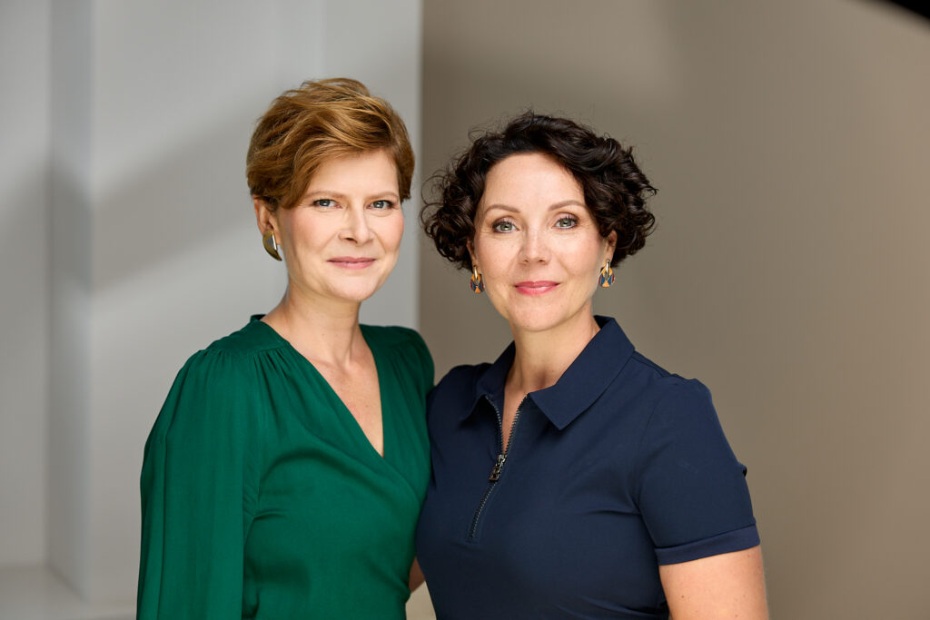 Na zdjęciu stoją obok siebie dwie kobiety patrzące w obiektyw z lekkim uśmiechem. Jedna ma krótkie blond włosy i zieloną sukienkę, druga krótkie ciemne kręcone włosy i granatową sukienkę z kołnierzykiem. Tło jest neutralne, co nadaje portretowi profesjonalny charakter.
