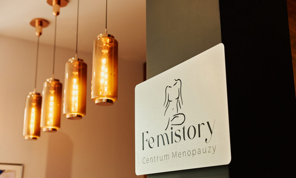 Na zdjęciu widać fragment wnętrza z eleganckimi, złocistymi lampami wiszącymi oraz tabliczką z logo „Femistory Centrum Menopauzy”. Logo przedstawia minimalistyczny zarys kobiecej sylwetki. Całość tworzy nowoczesną i stylową atmosferę.