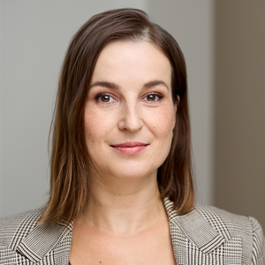 Kobieta o prostych brązowych włosach w marynarce w kratę patrzy w obiektyw.