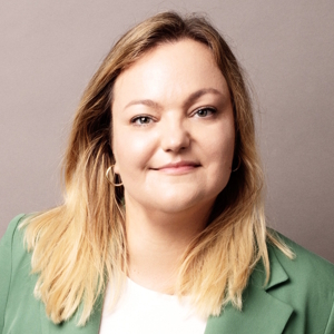 Kobieta o blond włosach w zielonej marynarce i białej bluzce patrzy w obiektyw.