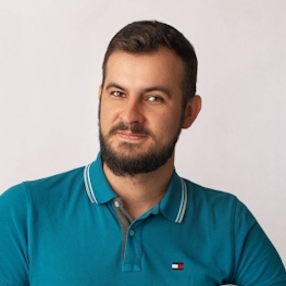 Mężczyzna z brodą w turkusowej koszulce polo uśmiecha się lekko do obiektywu.