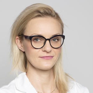 Kobieta w okularach o blond włosach, ubrana w białą koszulę, patrzy w obiektyw.