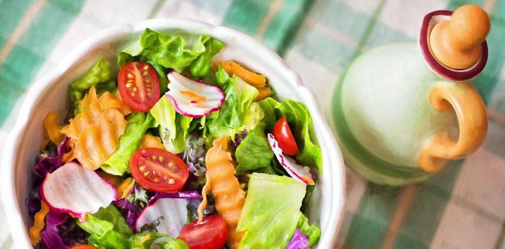 Na zdjęciu widać miskę pełną kolorowej sałatki z liśćmi sałaty, pomidorkami koktajlowymi, rzodkiewką, marchewką i kapustą. Obok stoi ceramiczna karafka, prawdopodobnie z oliwą. Całość ustawiona jest na obrusie w kratkę, tworząc świeży i apetyczny obraz.