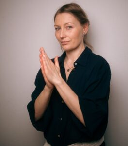 Na zdjęciu widać kobietę w granatowej koszuli stojącą na tle jasnej ściany. Ma związane włosy i delikatny uśmiech, a jej dłonie są złożone jakby w geście pocierania lub przygotowania. Wygląda spokojnie i skupiona.