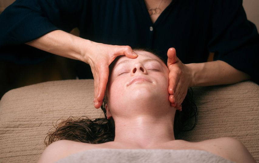 Na zdjęciu osoba leży z zamkniętymi oczami, a masażysta delikatnie masuje jej twarz, trzymając dłonie przy policzkach i czole. Twarz wygląda na zrelaksowaną i spokojną. Cała scena ma charakter relaksacyjny i kojący.