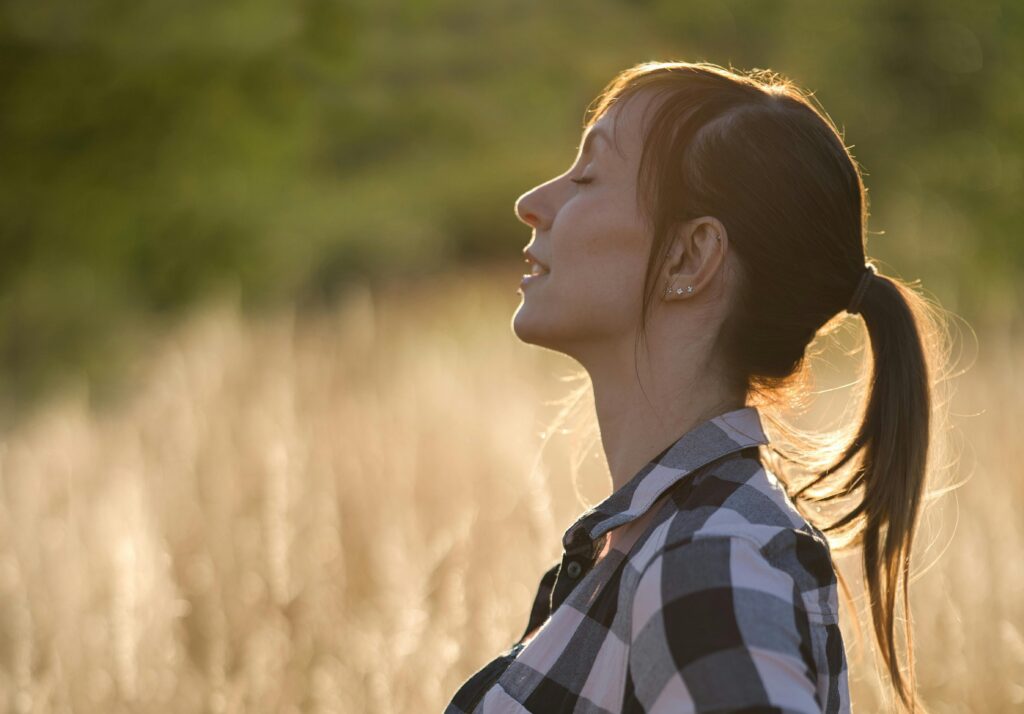 Na zdjęciu kobieta w koszuli w kratę stoi na tle pola w ciepłym świetle zachodzącego słońca. Ma związane włosy w kucyk i zamknięte oczy, a na twarzy wyraz spokoju i relaksu. Całość tworzy atmosferę wyciszenia i kontaktu z naturą.