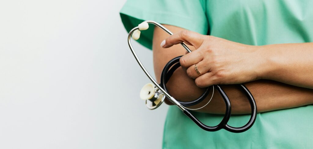 Na zdjęciu widać osobę w zielonym stroju medycznym trzymającą w ręku stetoskop. Ręce są skrzyżowane, a na palcu widoczna jest złota obrączka. Tło jest jasne i neutralne, co podkreśla profesjonalny charakter ujęcia.