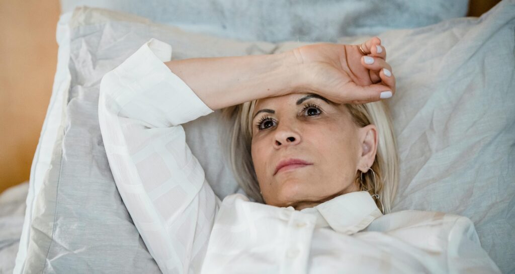 Na zdjęciu kobieta leży w łóżku, opierając głowę na poduszce i mając uniesione ramię na czole. Jej twarz wyraża zmęczenie lub zamyślenie. Ubrana jest w białą koszulę, a cała scena ma spokojny, ale nieco przygnębiony nastrój.