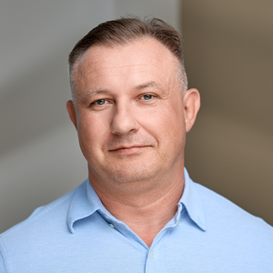 Mężczyzna w jasnoniebieskiej koszuli patrzy prosto w obiektyw.