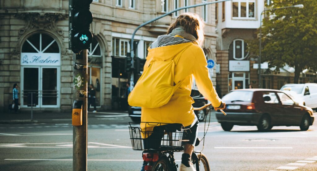 Na zdjęciu osoba w żółtej kurtce i plecaku jedzie na rowerze po mieście, zatrzymując się na skrzyżowaniu przy zielonym świetle dla rowerzystów. Na bagażniku roweru znajduje się koszyk. W tle widać budynki, samochody i przechodniów.