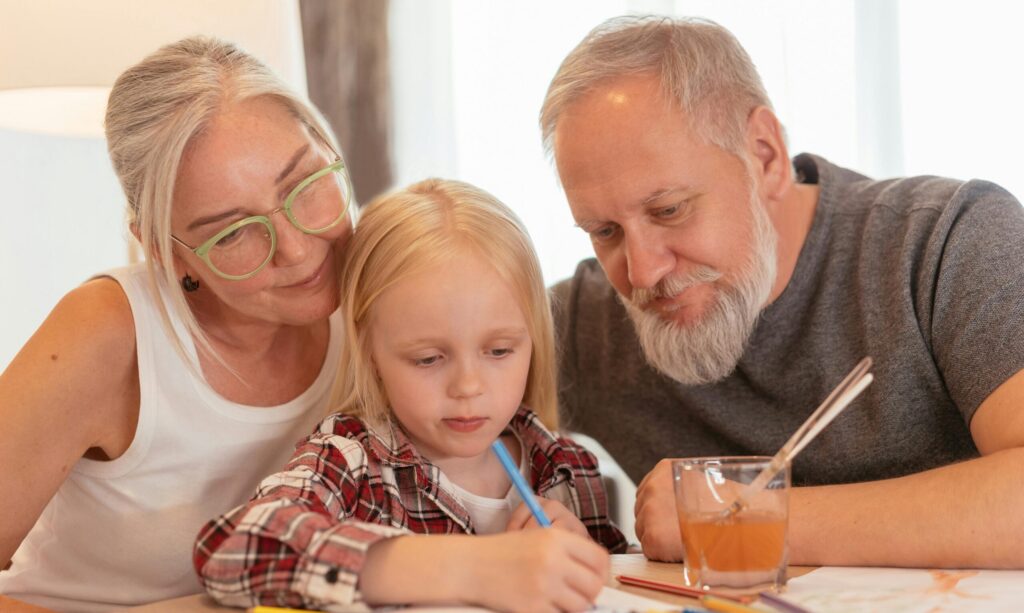 Na zdjęciu starsza kobieta i mężczyzna siedzą obok małej dziewczynki, która rysuje kolorowymi kredkami. Kobieta uśmiecha się ciepło, a mężczyzna z siwą brodą patrzy z zainteresowaniem na rysunek. Na stole leży szkic, kredki i szklanka z napojem.