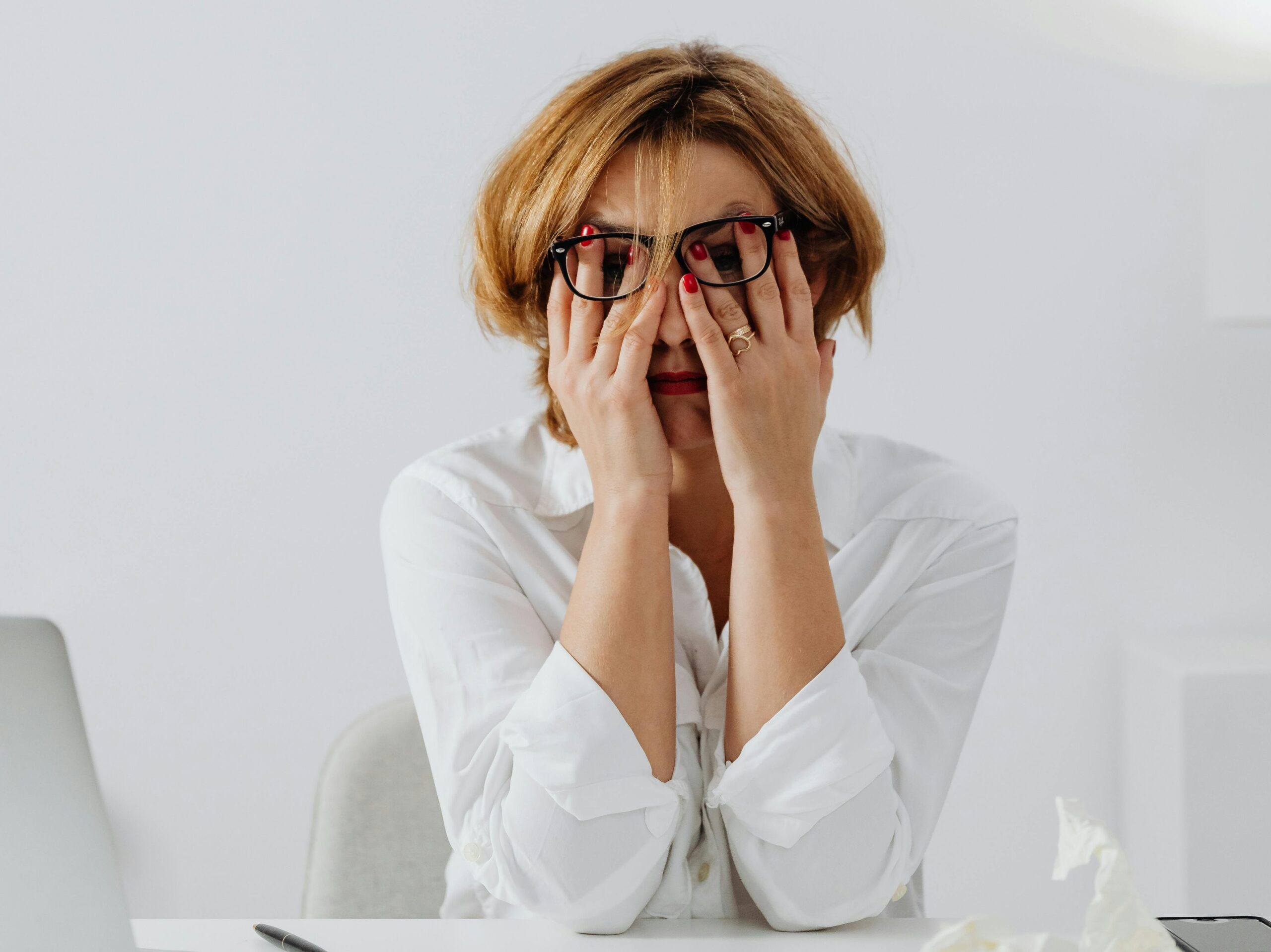 Kobieta w białej koszuli i okularach siedząca przy biurku, zasłaniająca dłońmi oczy i czoło, wygląda na zmęczoną lub zestresowaną.
