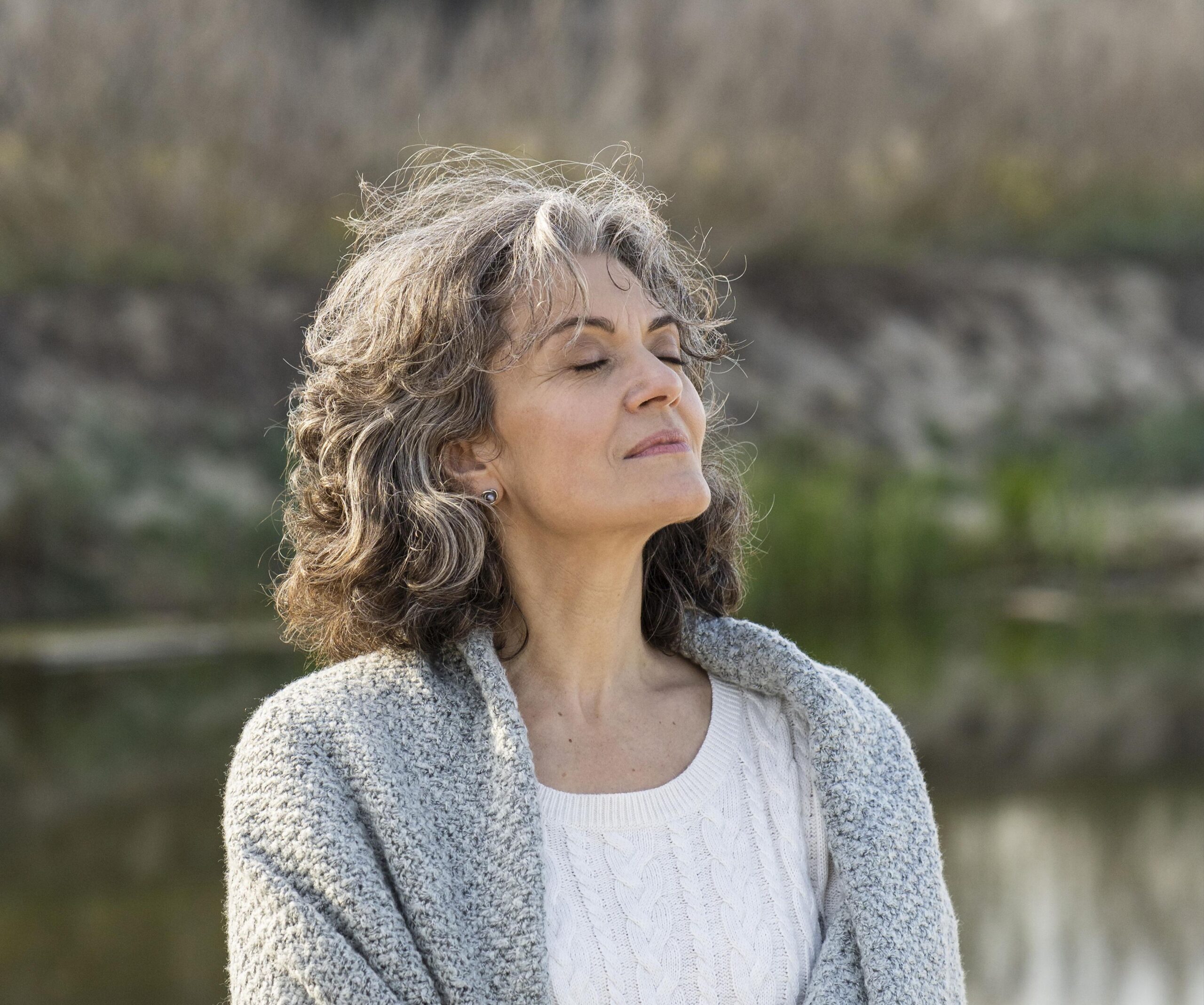 Zbliżenie w plenerze, na którym widać kobietę z kręconymi, siwiejącymi włosami, która ma zamknięte oczy i uniesioną lekko głowę, jakby wchłaniała słońce lub świeże powietrze. Ma na sobie biały sweter z warkoczowym splotem oraz szary, narzucony na ramiona, dzianinowy szal lub kardigan. W tle widać niewyraźny brzeg wody (może jeziora lub stawu) oraz brunatną, rozmytą roślinność. Oświetlenie jest naturalne i jasne.