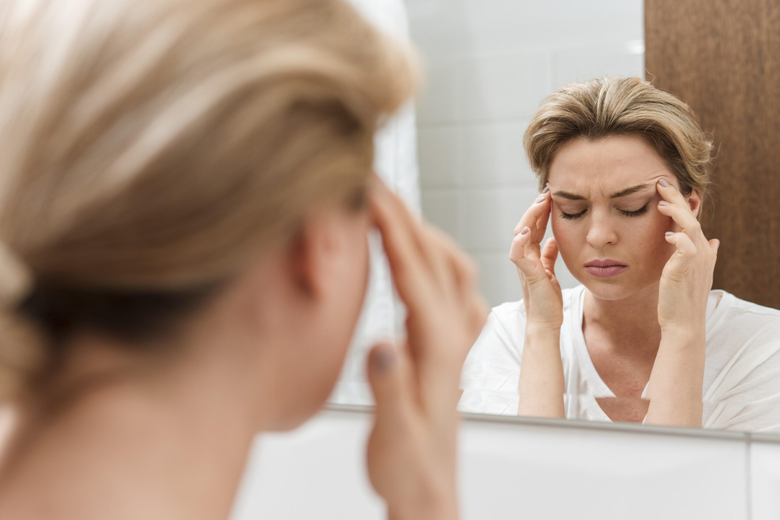 Kobieta z jasnymi włosami, ubrana w białą koszulkę, stoi przed lustrem w łazience, trzymając obie dłonie przy skroniach. Jej oczy są zamknięte, a twarz jest wyraźnie ściągnięta w grymasie bólu, co sugeruje, że cierpi na silny ból głowy lub migrenę. Odbicie w lustrze, ostre i skupione, kontrastuje z lekko rozmytym obrazem jej tyłu głowy i ręki na pierwszym planie.