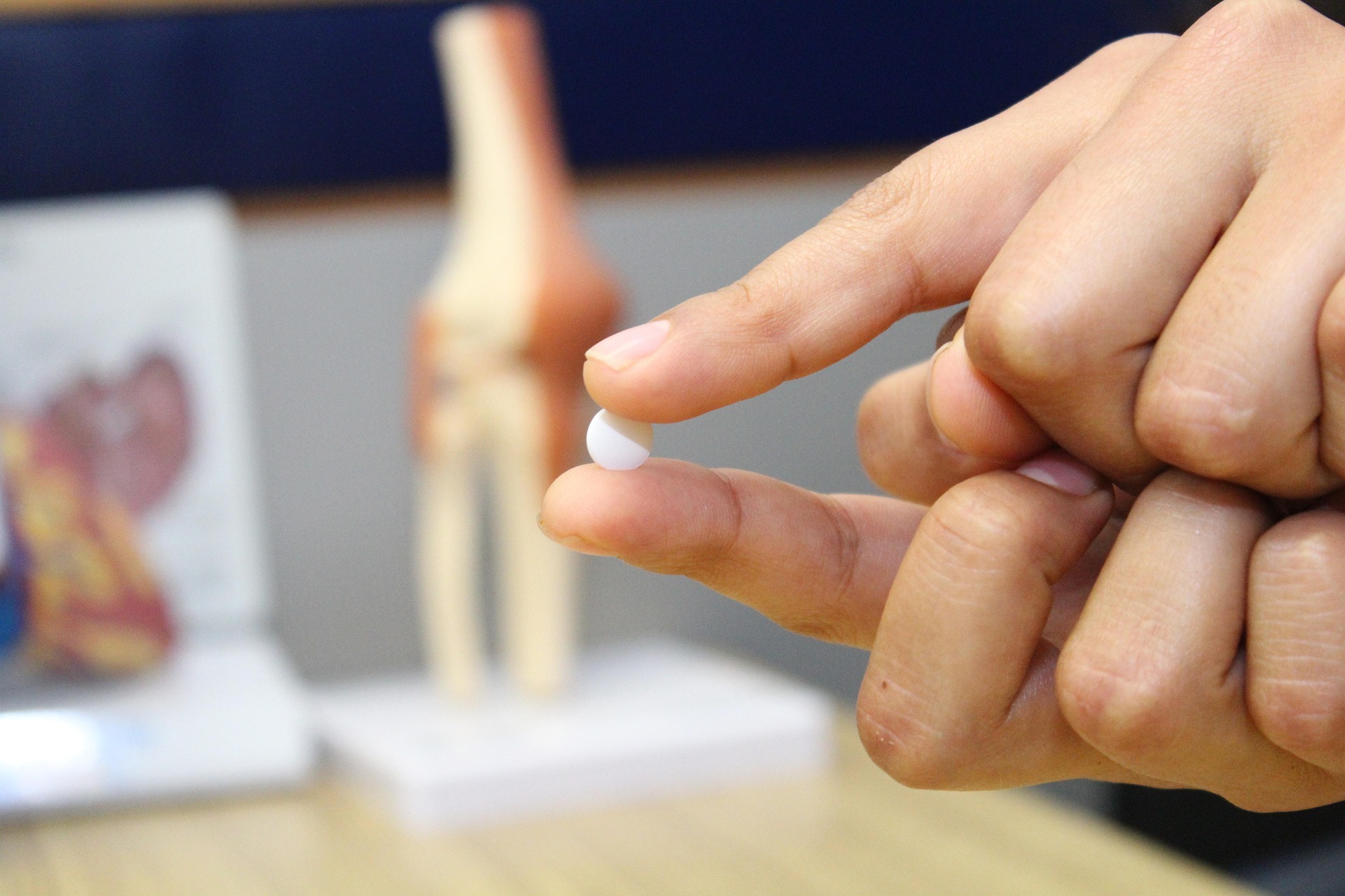 Zbliżenie na rękę trzymającą małą, okrągłą, białą tabletkę między kciukiem a palcem wskazującym. Tło jest rozmyte, ale widać na nim model anatomiczny stawu kolanowego oraz inny model ciała ludzkiego po lewej. Ręka i tabletka są w ostrym ujęciu na pierwszym planie.