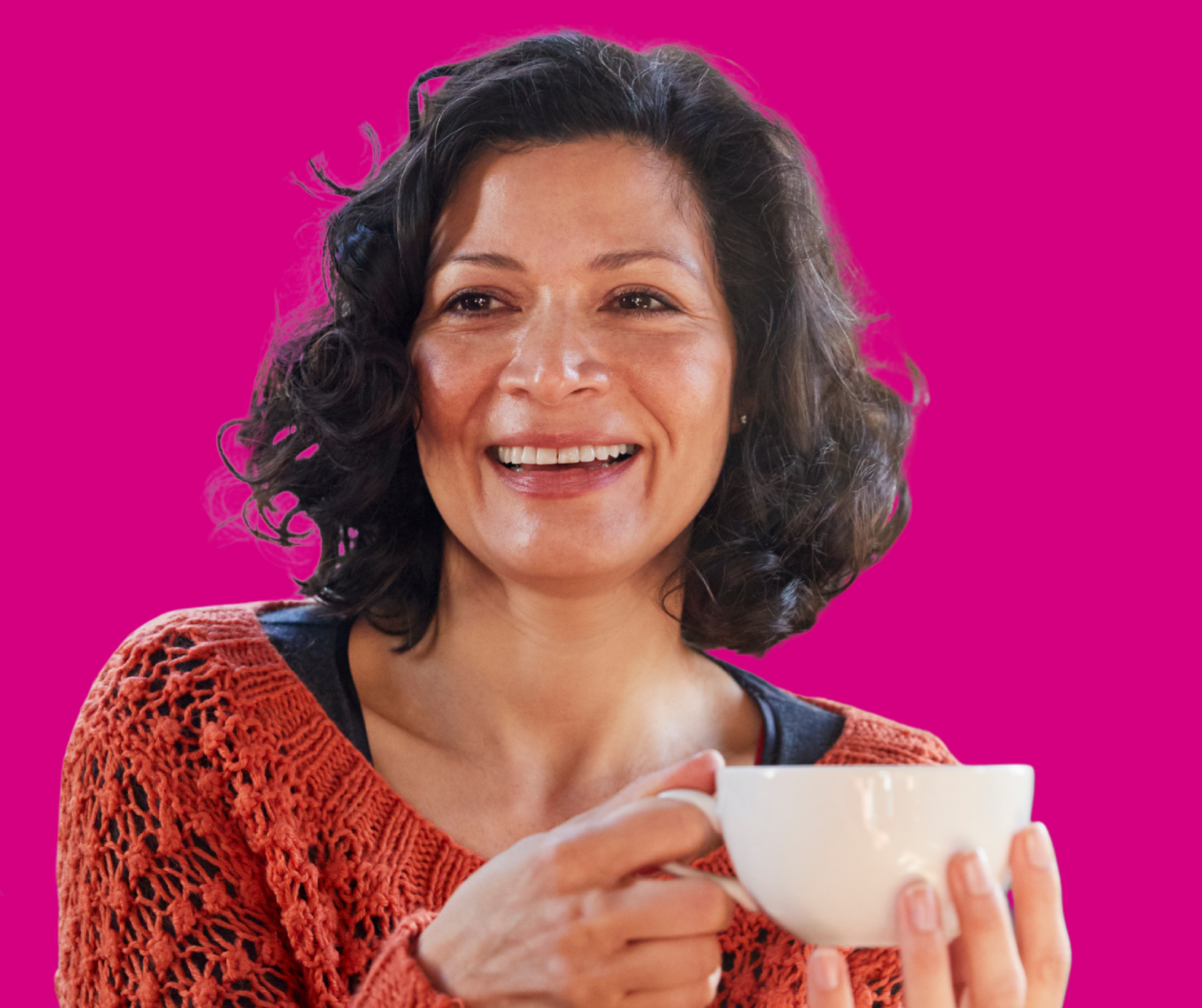 Zdjęcie przedstawia radosną kobietę w wieku średnim, patrzącą lekko w bok z szerokim uśmiechem. Ma ciemne, sięgające do ramion kręcone włosy. Ubrana jest w pomarańczowy, ażurowy sweter o grubym splocie, pod którym widoczna jest ciemna bluzka. Kobieta trzyma oburącz białą, ceramiczną filiżankę, jakby przygotowywała się do wypicia napoju. Tło jest jednolite, w jaskrawym kolorze fuksji (intensywny róż), co mocno kontrastuje z postacią.