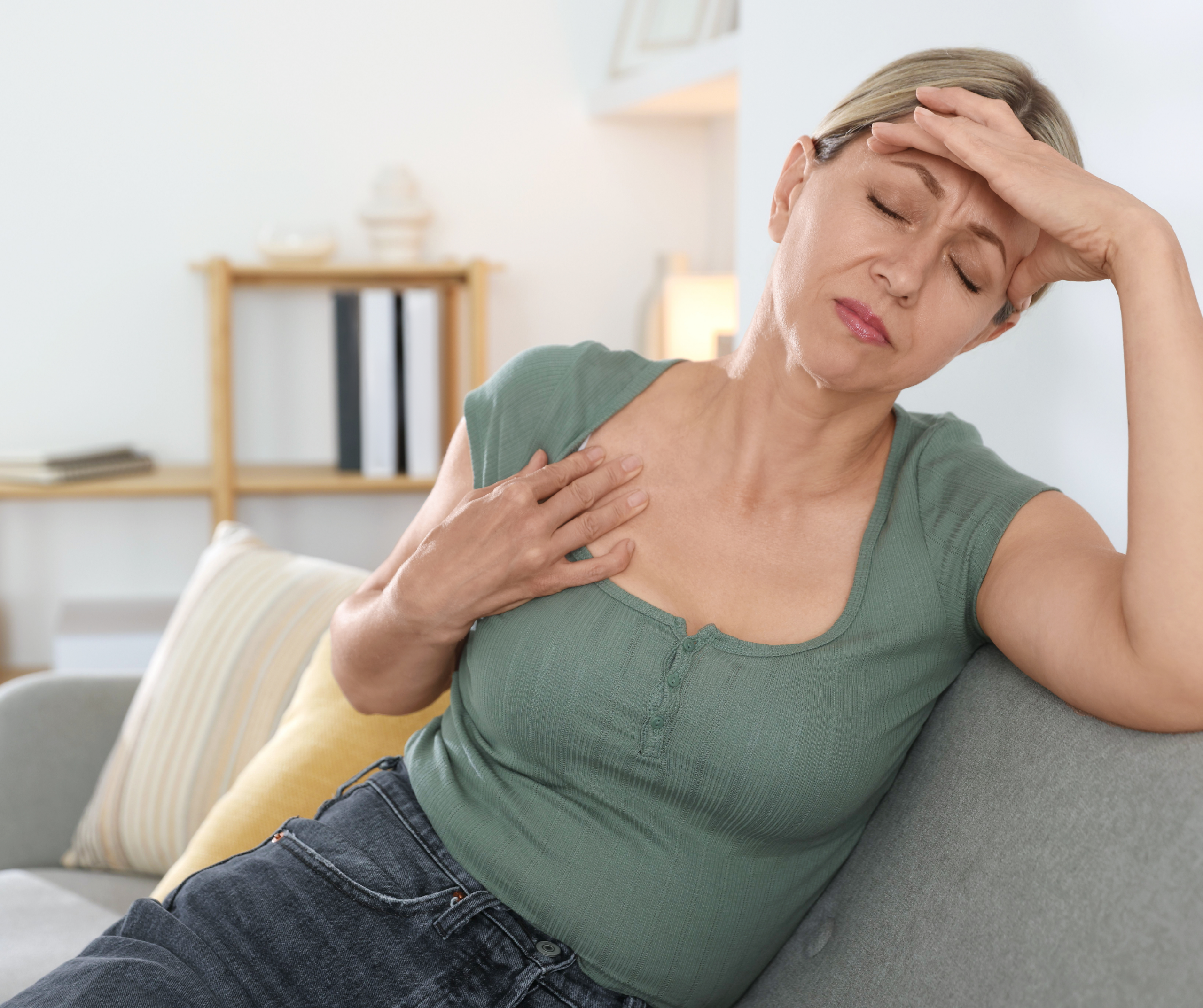 Zdjęcie przedstawia dojrzałą kobietę siedzącą na szarej kanapie w jasnym, domowym wnętrzu. Kobieta ma przymknięte oczy i wyraz twarzy sugerujący dyskomfort lub złe samopoczucie. Jedną dłoń trzyma przyłożoną do czoła, a drugą opiera na klatce piersiowej, tuż pod dekoltem. Ubrana jest w zieloną bluzkę z krótkim rękawem i ciemne spodnie. W tle widać rozmyte wnętrze salonu z drewnianym regałem, na którym stoją książki i dekoracje, co tworzy spokojną, ale sugestywną atmosferę związaną z domowym zaciszem i kwestiami zdrowotnymi.