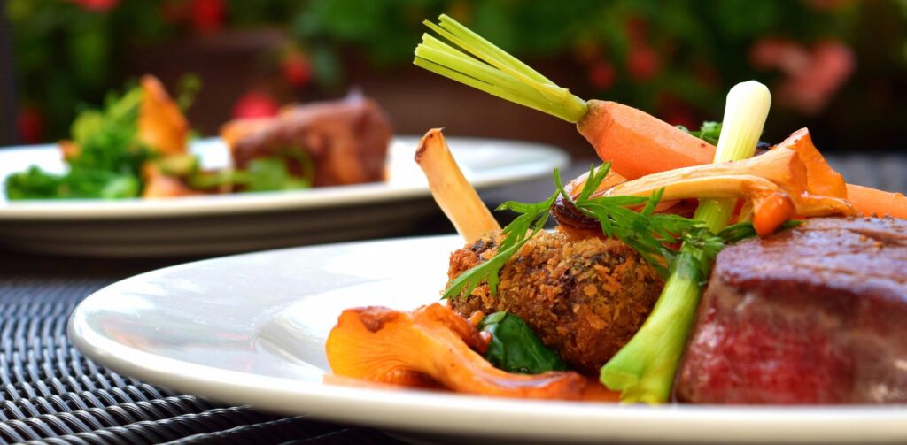 Zbliżenie pod kątem na elegancko zaserwowany posiłek na białym, płytkim talerzu. Talerz stoi na ciemnym, plecionym stole, a w rozmytym tle widać drugą porcję oraz ogród z czerwonymi kwiatami. Szczegóły potrawy: Główny element: Soczysty, gruby kawałek pieczonego mięsa (prawdopodobnie polędwica wołowa) o rumianej skórce. Dodatki: * Złociste, całe kurki (grzyby) o charakterystycznym kształcie. Chrupiący, panierowany element (krokiet lub kotlecik). Młode, mini marchewki z pozostawionymi zielonymi końcówkami. Świeży szczypiorek oraz natka pietruszki dekorująca wierzch dania. Stylistyka: Zdjęcie charakteryzuje się płytką głębią ostrości (bokeh), co skupia całą uwagę na teksturach jedzenia w pierwszym planie. Kolory są nasycone i naturalne.