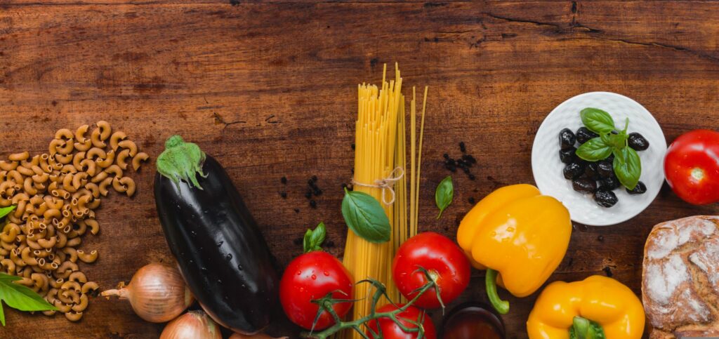 Widok z góry (flat lay) na bogatą kompozycję świeżych warzyw i produktów spożywczych, ułożonych w dolnej części kadru na tle ciemnego, rustykalnego drewnianego blatu. Górna część zdjęcia pozostaje pusta, tworząc idealną przestrzeń na napis lub logo. Co znajduje się na zdjęciu: Warzywa: Dorodny, lśniący bakłażan, soczyste czerwone pomidory na gałązce, dwie żółte papryki, cebule oraz świeże liście bazylii. Produkty sypkie i makarony: Pęczek długiego spaghetti przewiązany sznurkiem oraz rozsypany obok ciemny makaron typu kolanka (prawdopodobnie pełnoziarnisty). Dodatki: Mała biała miseczka z czarnymi oliwkami udekorowanymi bazylią, bochenek świeżego chleba oprószony mąką oraz rozsypane ziarenka czarnego pieprzu.